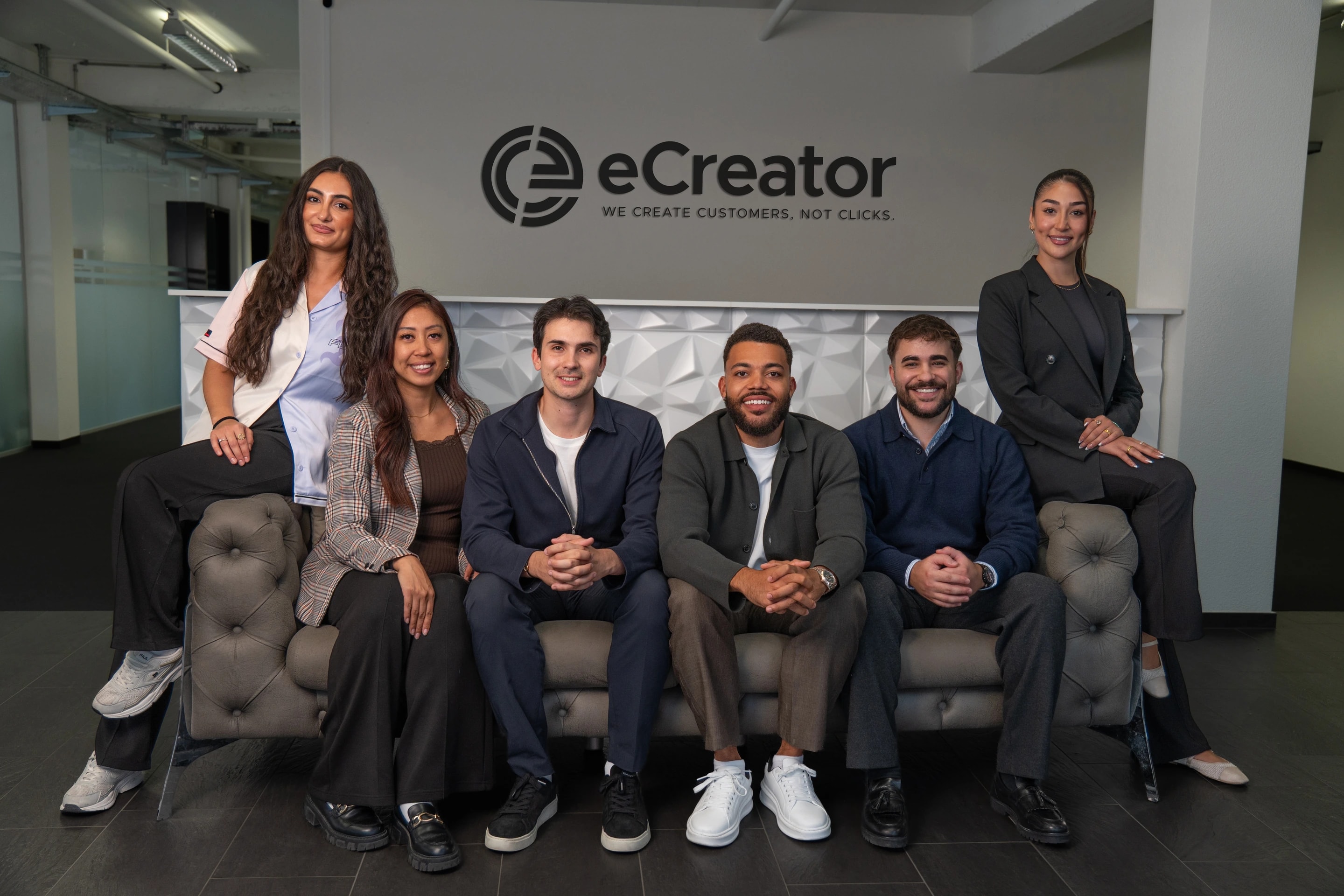 Das Team von eCreator im Büro: sechs Personen auf und neben einem Sofa vor der Wand mit dem eCreator-Logo